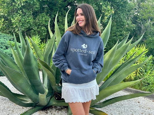 A woman wearing a slate blue hoodie with a white 'spotted seal NORFOLK' logo stands outdoors in front of a large agave plant. The hoodie features a white seal logo and the text 'spotted seal NORTHEM' printed on the chest.