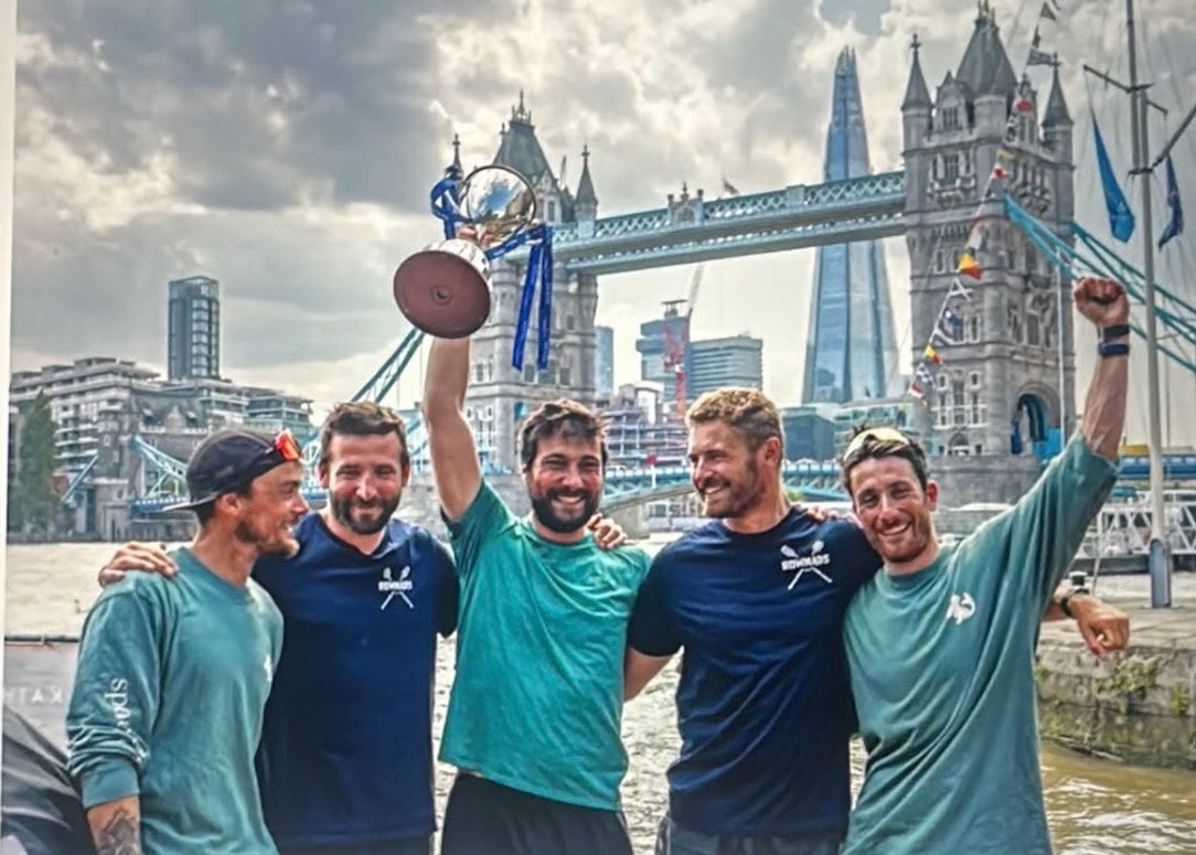 Five men celebrating on a riverbank with Tower Bridge and London skyline in the background, with one man holding up a trophy. They are wearing spotted-seal long sleeve tees
