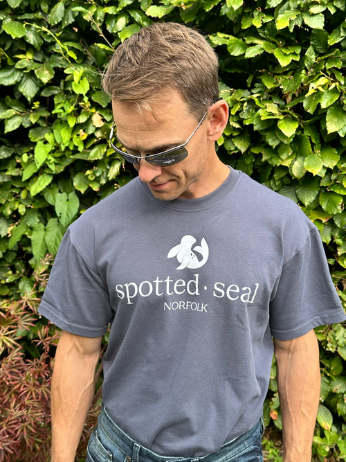 man wearing a blue-grey t-shirt with spotted-seal logo standing among green foliage.