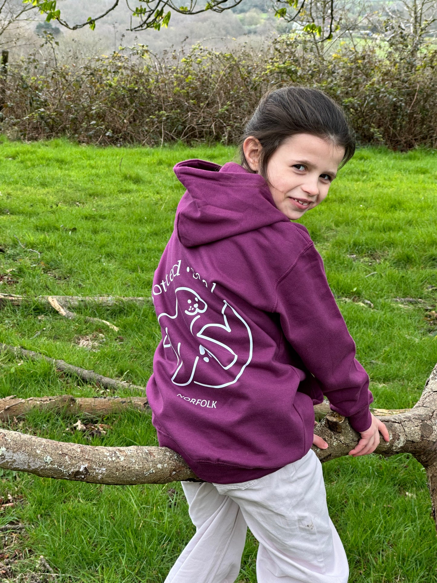 Child in a purple hoodie standing next to a tree in a grassy area
