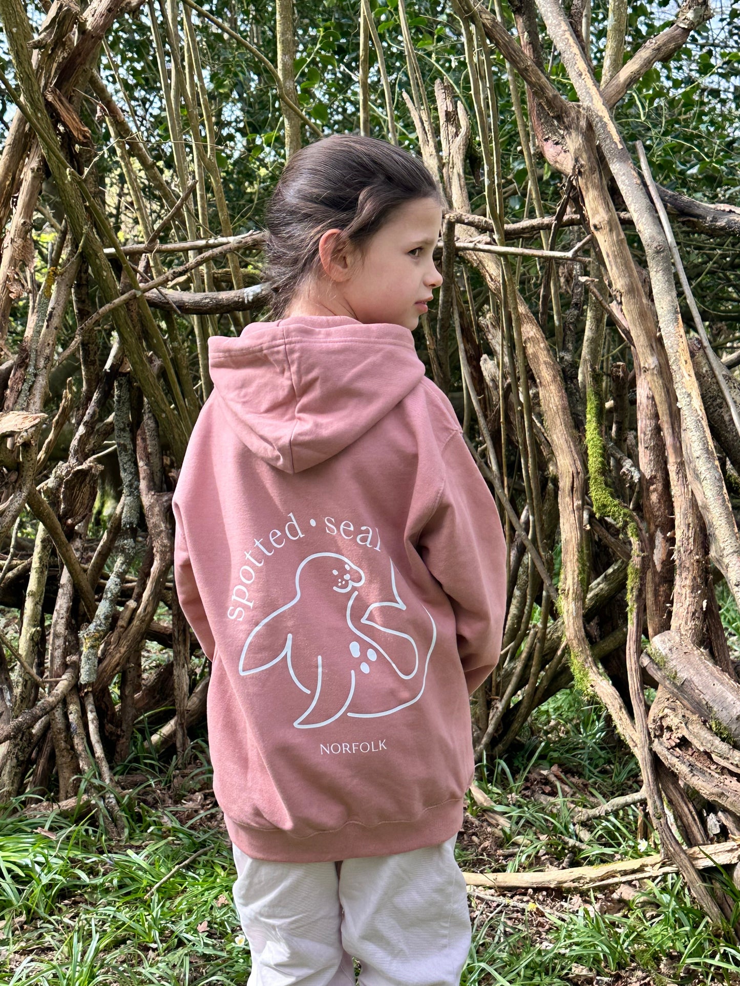Person wearing a pink hoodie with a logo standing among stacked wooden logs outdoors.
