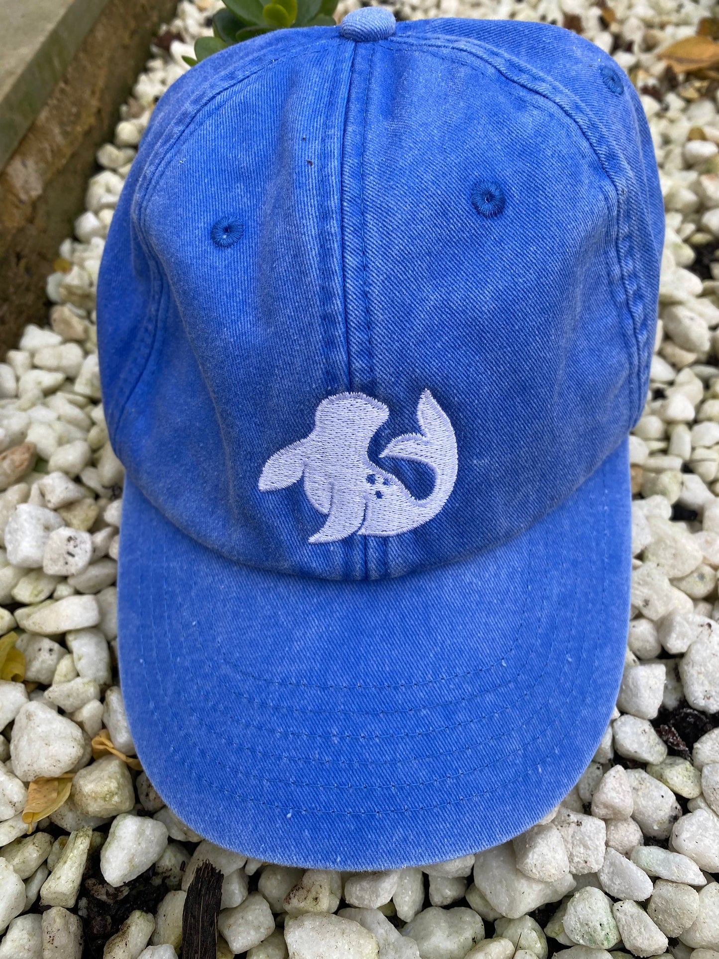 spotted-seal unisex cap in cornflower blue Stiffkey skies colour on white stones with embroidered seal